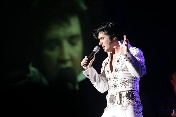 Ein Sänger in einem weißen, mit goldenen Nieten verzierten Elvis-Kostüm singt auf einer Bühne ins Mikrofon.